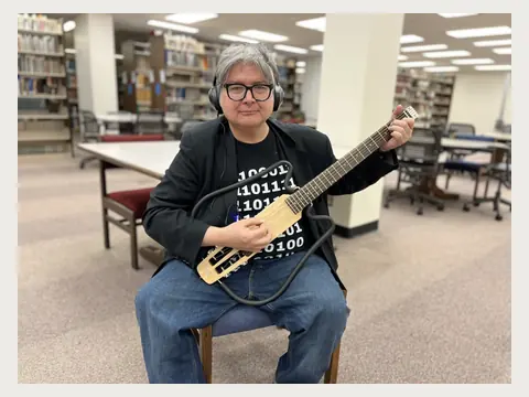 Herbert Midgley playing a compact electric guitar in a library.